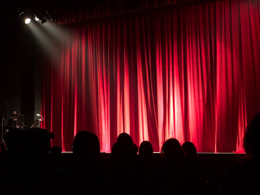 Dimly lit theater stage with red curtains and audience silhouettes under spotlights